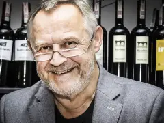 Marek Kondrat, z uśmiechem i okularami, na tle butelek wina. Jego majątek to nie tylko sukcesy zawodowe, ale i pasja do trunków.