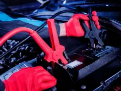 Mechanik w czerwonych rękawiczkach podłącza kable rozruchowe do akumulatora. Czy nowy akumulator trzeba ładować? Sprawdźmy!
