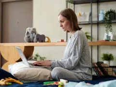 Kobieta w piżamie pisze na laptopie, przygotowując wniosek o urlop macierzyński. Obok pluszowy królik i zabawki.