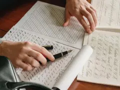 Manos practicando caligrafía con una pluma sobre hojas con ejercicios de letras.