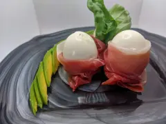 Due uova di quaglia sode avvolte in prosciutto crudo, servite con fette di avocado e foglie verdi su un piatto scuro.