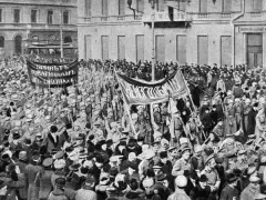 Multitud con pancartas y uniformes militares marchando en una calle, con edificios al fondo.