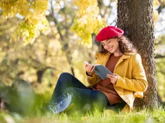 Mujer con boina roja lee un libro bajo un árbol, inspirada por los temas de cuentos del otoño.