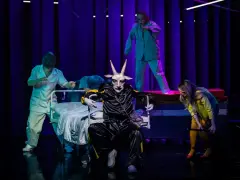 Na scenie, w surrealistycznej atmosferze, panna obsada w stroju z głową kozła siedzi na wózku. Wokół niej, w jasnych fartuchach, poruszają się postacie przypominające personel medyczny.