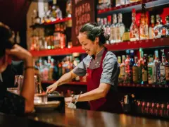 Uśmiechnięta barmanka przygotowuje drinki za barem pełnym alkoholi. Zastanawiasz się, ile zarabia barman w restauracji?