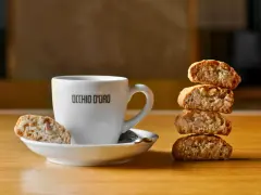 Ein weißer Kaffeebecher mit "OCCHIO D'ORO" Schriftzug steht neben einem Stapel Cantuccini. Ein Cantuccino liegt auf der Untertasse. Dieses Bild inspiriert zum besten Cantuccini Rezept.