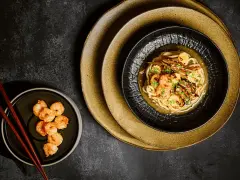 Chintan ramen z krewetkami i warzywami, podany na czarnym talerzu, obok miseczki z dodatkowymi krewetkami i pałeczkami.