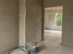 Wnętrze w trakcie prac wykończeniowych, ściany pokryte tynkami gipsowymi lub cementowo wapiennymi. Widoczne wiadro z zaprawą i narzędzia.