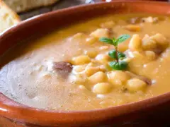 Una ciotola di terracotta piena di zuppa di fagioli cremosa, con pezzetti di carne e una fogliolina di menta. Perfetta per una cena confortante.