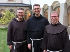 Drei Franziskaner Minoriten in braunen und schwarzen Kutten lächeln in die Kamera vor einer Klosterfassade.