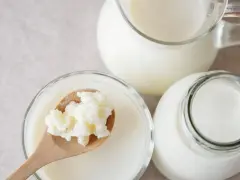 Ziarna kefirowe na łyżce nad miseczką z maślanką i butelką z kefirem.