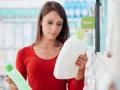 Mujer compara dos botellas de detergente, leyendo la etiqueta producto de limpieza.