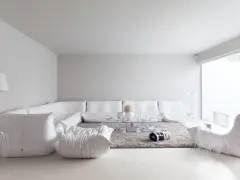 Soggiorno minimalista con divano componibile bianco, tappeto grigio e tavolino trasparente. Le tonalità di bianco per pareti creano un'atmosfera eterea.