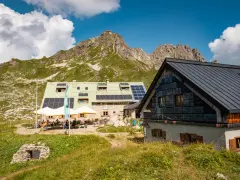 Die Buchel Alpe, eine Berghütte mit Solaranlagen, liegt vor einer beeindruckenden Bergkulisse. Gäste sitzen draußen unter Sonnenschirmen.