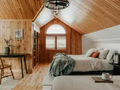 Dormitorio con paredes y techo de madera, cama con mantas y almohadas, ventana arqueada y escritorio.