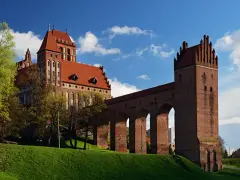 Majestatyczny zamek w Kwidzynie, z czerwonej cegły, z charakterystycznym gdaniskiem i długim, arkadowym murem obronnym, wznosi się na zielonym wzgórzu pod błękitnym niebem.