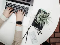 Kobieta pracuje na laptopie, obok leżą okulary, telefon i roślina. To idealne zdjęcia w chmurze do inspiracji.