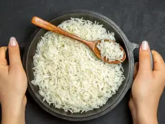Dwie ręce trzymają okrągłą, czarną patelnię pełną puszystego ryżu basmati. Drewniana łyżka zanurzona w ryżu.