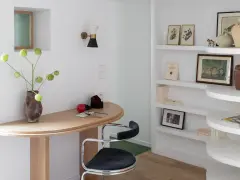 Un despacho pequeño en casa con un escritorio curvo de madera, una silla de terciopelo azul y estanterías blancas llenas de libros y objetos decorativos.