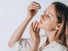 Frauenhand trägt Tropfen eines Serums auf ihr Gesicht auf. Ob Squalan oder Hyaluron, die Haut strahlt Feuchtigkeit aus.