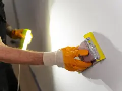 Ręka w pomarańczowej rękawicy wykonuje szlifowanie tynku gipsowego za pomocą gąbki ściernej.
