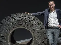 Wim Delvoye pose à côté d'une roue ornée de dragons, une œuvre d'art monumentale.
