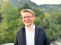 Ein Mann mit Brille lächelt in die Kamera. Er trägt ein weißes Hemd und eine dunkle Jacke. Im Hintergrund sind Bäume und Berge zu sehen. Er studiert Seelsorge.