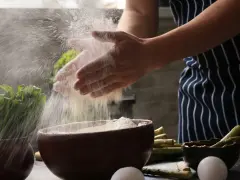 Hände verarbeiten Maniokmehl, das in der Luft schwebt. Daneben liegen Spargel und Eier, bereit für die Zubereitung.