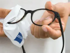 Dłonie czyszczą okulary okularowe białą ściereczką z logo Zeiss.