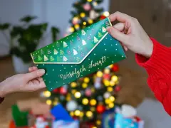 Jak zapakować voucher na prezent? Zielona koperta z choinkami i napisem "Wesołych Świąt!" jest przekazywana w świątecznej atmosferze.