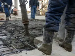 Beton na fundamenty wylewany jest na zbrojenie. Pracownicy w gumowcach nadzorują proces.