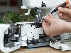 Dłonie regulują maszynę do szycia, używając śrubokręta. Widać fragmenty mechanizmu i nitki, a także pasek napędowy.