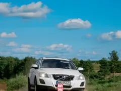 Białe Volvo XC60 T6 jedzie po piaszczystej drodze wśród zieleni, pod błękitnym niebem z chmurami.