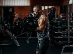 Mężczyzna ćwiczy biceps z ciężarem. W tle siłownia, sprzęt i inny ćwiczący.