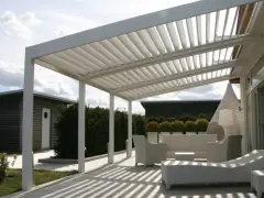 Un moderno porche de obra con techo de lamas ajustables, mobiliario de exterior blanco y un camino de madera que conduce a una cabaña.