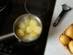 Ziemniaki gotują się w garnku na płycie indukcyjnej. Obok leży obieraczka i koszyk ze świeżymi ziemniakami. Oto jak ugotować ziemniaki.