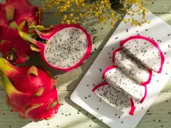 Dwa całe i połówka owocu pitaya, obok pokrojone w ćwiartki na białej desce.