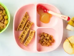Baby-freundliche Apfelwaffeln mit Apfelmus und Zimt-Apfelstückchen auf einem rosa Teller. Ein halber Apfel und eine Banane liegen daneben.