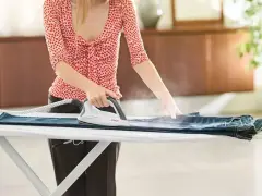 Donna stira un paio di jeans su un asse da stiro, usando un ferro da stiro a vapore. Impara come stendere i pantaloni per un look impeccabile.