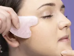 Eine Hand nutzt ein Gua Sha Stein für eine Gesichtsmassage. Das Werkzeug ist herzförmig und aus rosafarbenem Stein.