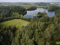 Malownicze jezioro Jaczno otoczone gęstym lasem, z małą wyspą pośrodku. W oddali widać pagórkowaty krajobraz pól i kolejnych lasów.