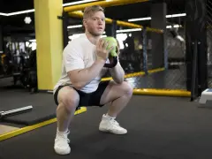 Mężczyzna w białej koszulce wykonuje przysiad z zielonym kettlebell, który przypomina kształtem puchar.