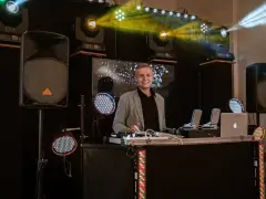 Uśmiechnięty dj na 18. urodziny, gotowy do rozkręcenia imprezy. W tle kolorowe światła i sprzęt nagłośnieniowy.