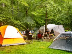 Grupa przyjaciół odpoczywa na polu namiotowym Mazury. Wokół rozstawione są namioty, a na stole piknikowym stoją napoje i przekąski.