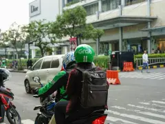Zwei Personen auf einem Roller, der Fahrer trägt einen grünen Helm mit dem "Grab" Logo. Sie sind unterwegs in Thailand, um mit der Grab App Kunden zu transportieren.