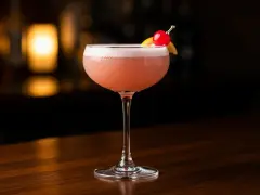 En rosa drink, en Pink Lady, med skum och garnering av körsbär och citronklyfta, står på en träyta.