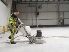 Pracownik w kamizelce odblaskowej wykonuje szlifowanie betonu za pomocą maszyny.