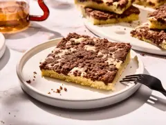Ein Stück versunkener Streuselkuchen mit Schokostreuseln auf einem Teller, daneben eine Gabel und ein Glas Tee.