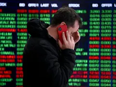 Hombre preocupado habla por teléfono frente a pantalla de bolsa de valores de Nueva York, con números rojos y verdes indicando un posible crack.