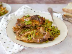 Un piatto di ossobuco con piselli e carote, servito su un piatto bianco con una forchetta. Sullo sfondo, fette di pane.